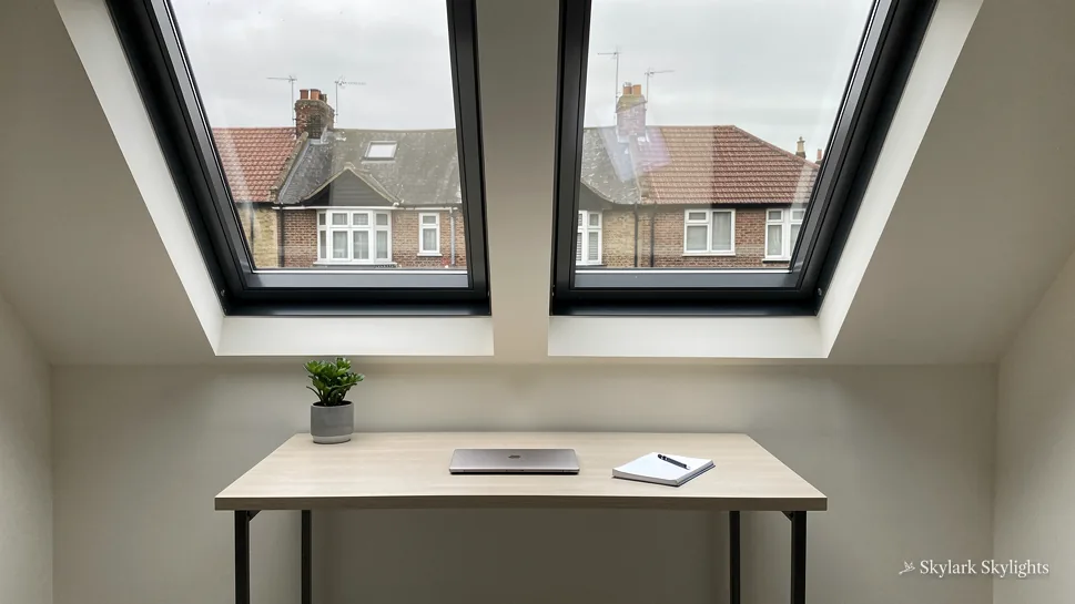 Desk below two dark-framed roof windows looking out over neighbouring roofs